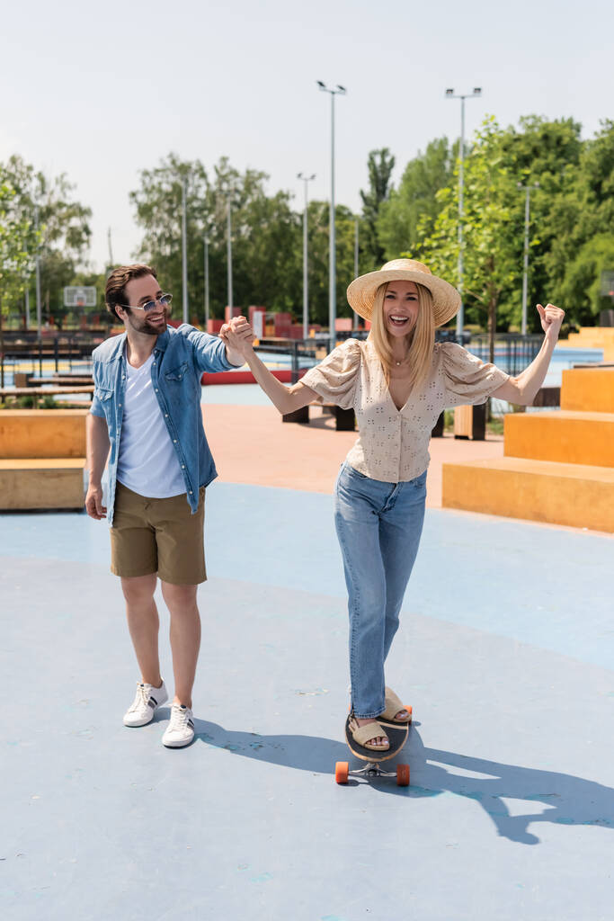 Excited woman in straw hat riding longboard and holding hand of boyfriend in skate park  - Photo, Image