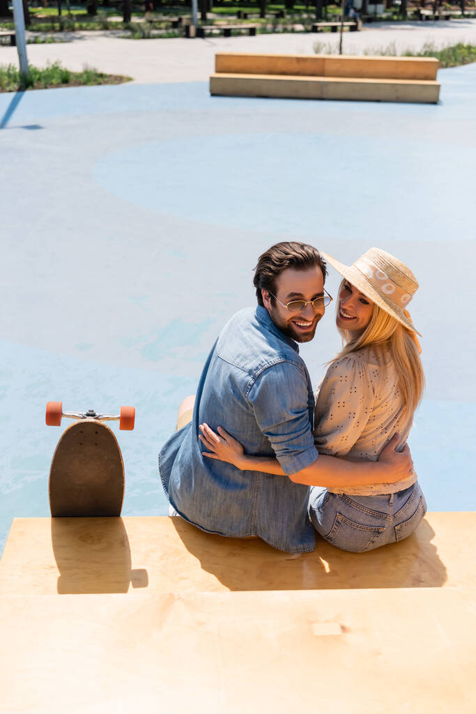 Улыбающаяся пара в соломенной шляпе и солнцезащитных очках, обнимающая длинную доску в скейт-парке  - Фото, изображение