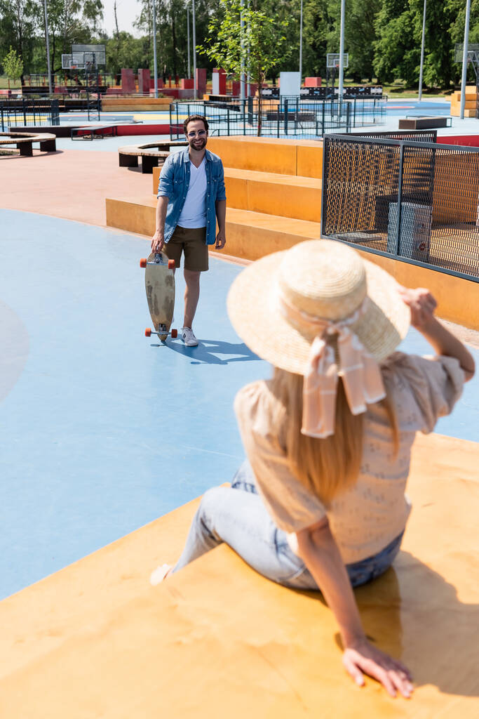Uomo sorridente in possesso di longboard vicino fidanzata offuscata in cappello di paglia in skate park  - Foto, immagini