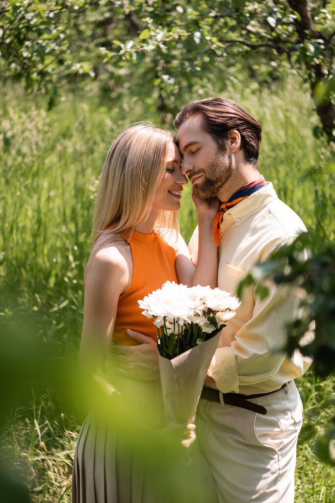 Vista laterale della donna sorridente che tiene il bouquet e tocca il fidanzato elegante nel parco  - Foto, immagini