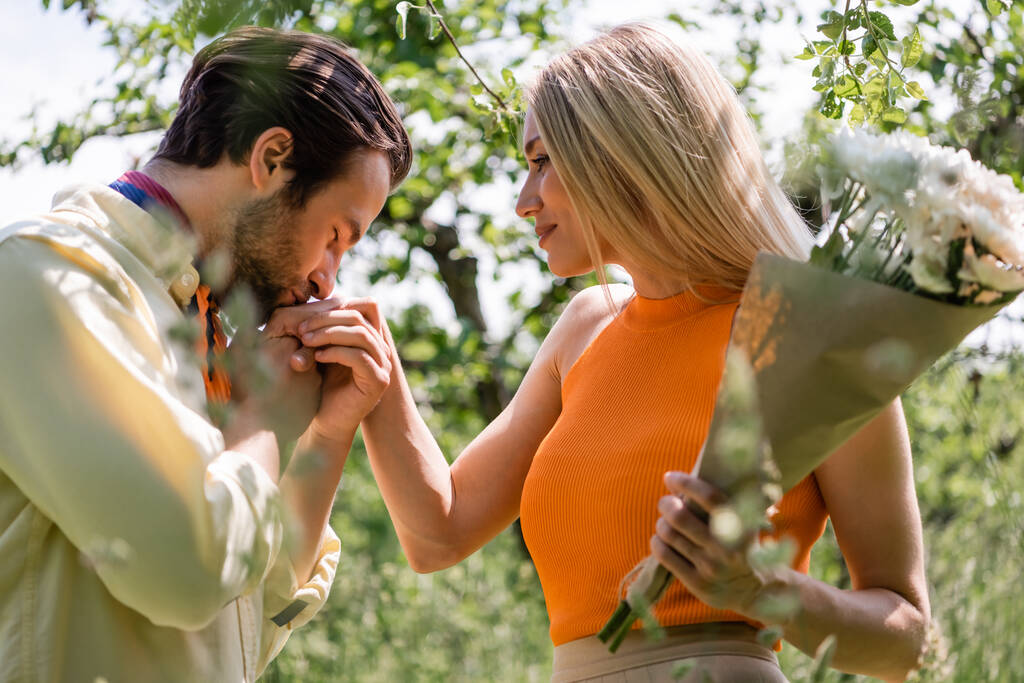Boční pohled na stylový muž líbání ruku přítelkyně držící kytici v parku  - Fotografie, Obrázek