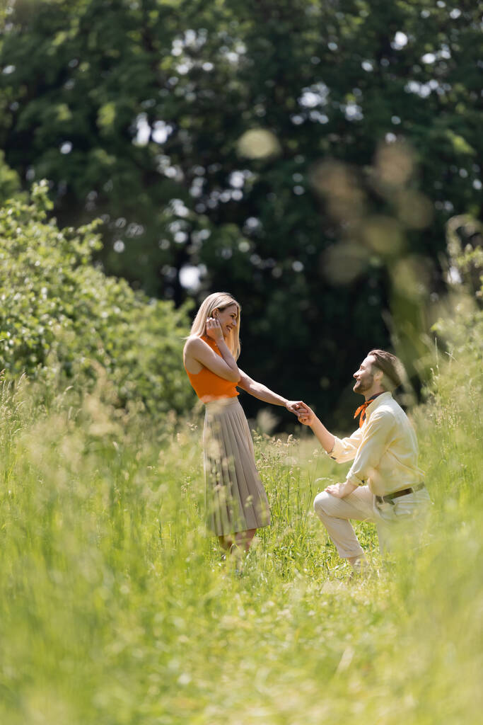 Widok z boku modny mężczyzna trzymając rękę dziewczyny i klęcząc na trawniku w parku  - Zdjęcie, obraz