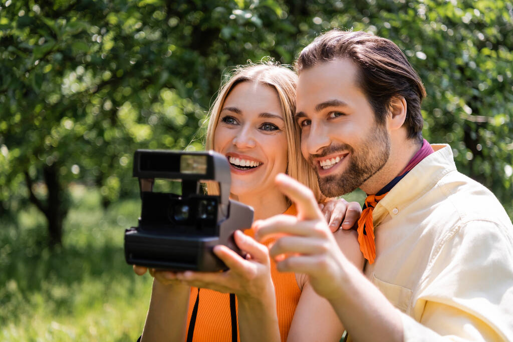 Homem elegante apontando com o dedo perto da namorada com câmera retro borrada no parque  - Foto, Imagem