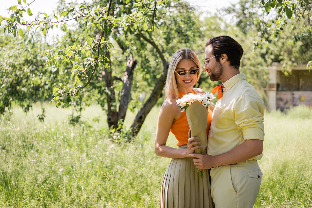 Homme à la mode dans les lunettes de soleil tenant bouquet près de petite amie souriante dans le parc d'été  - Photo, image