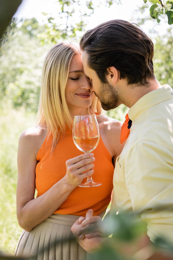 Smiling woman holding glass of wine and hand of stylish boyfriend in summer park  - Photo, Image