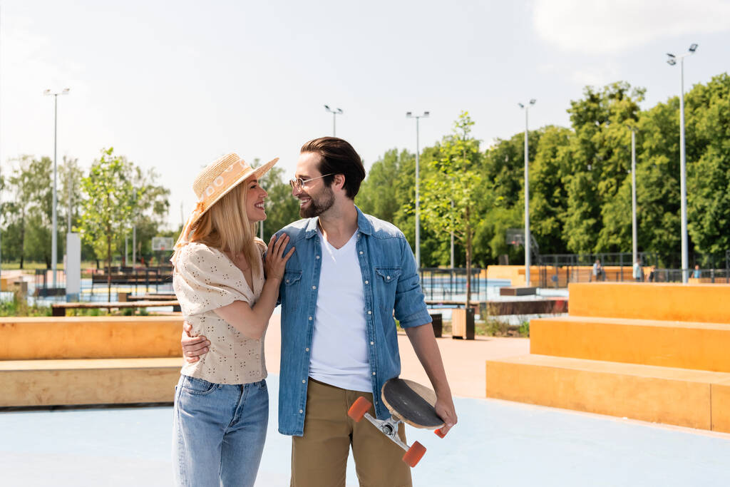 Вид сбоку улыбающейся женщины, обнимающей молодого парня с лонгбордом в скейт-парке  - Фото, изображение