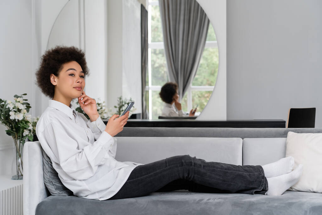 mujer afroamericana soñadora sosteniendo teléfono inteligente mientras descansa en un sofá de terciopelo en la sala de estar moderna  - Foto, imagen