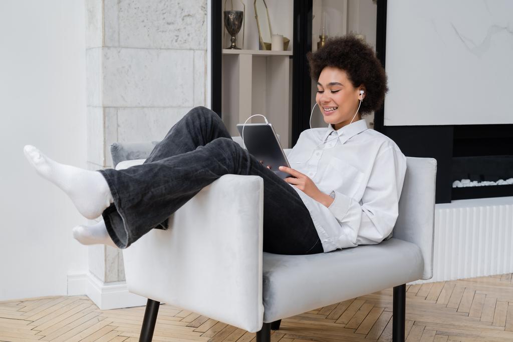 felice donna afroamericana in auricolare con tablet digitale mentre guarda film e seduto sulla poltrona grigia  - Foto, immagini