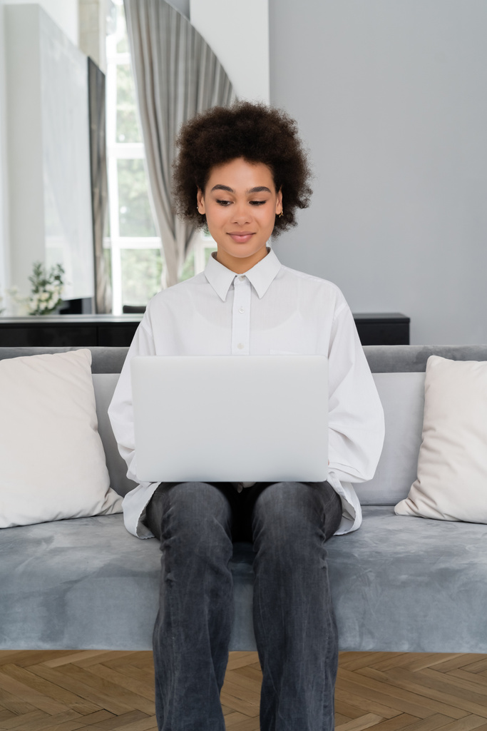 усміхнена афроамериканська жінка використовує ноутбук, сидячи на сірому оксамитовому дивані
 - Фото, зображення