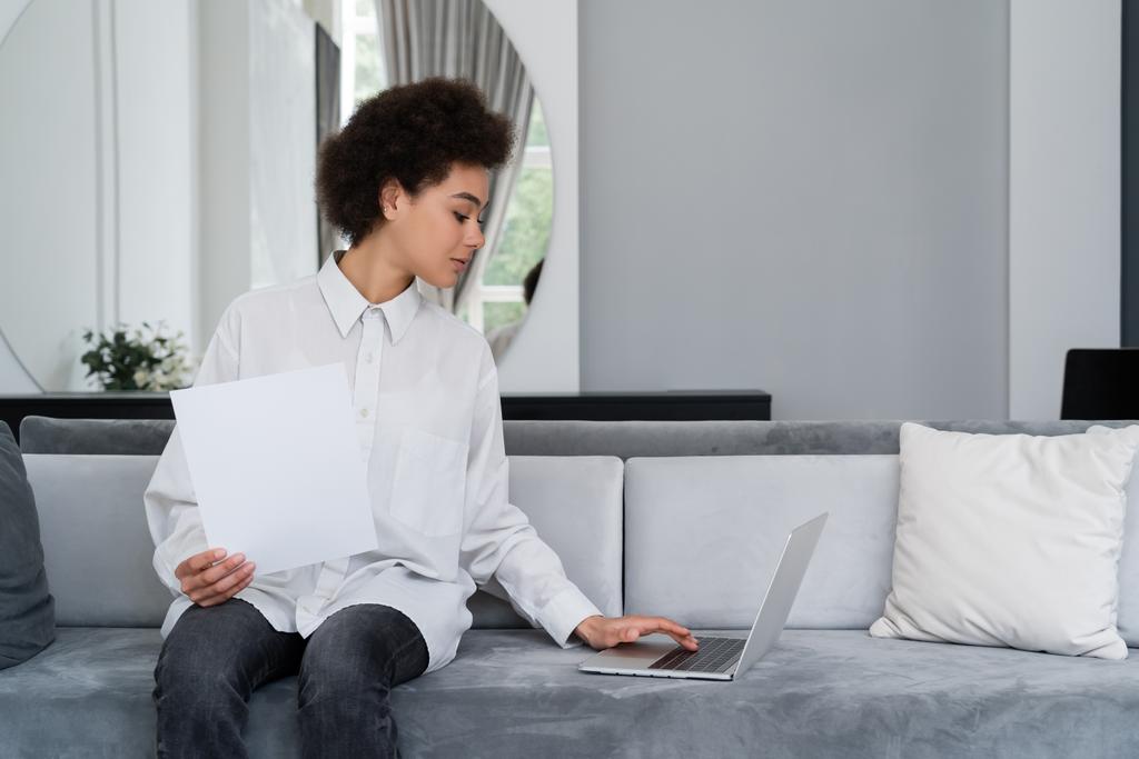 Afrikkalainen amerikkalainen nainen katselee tyhjä asiakirja ja käyttää kannettavaa tietokonetta sametti harmaa sohva - Valokuva, kuva