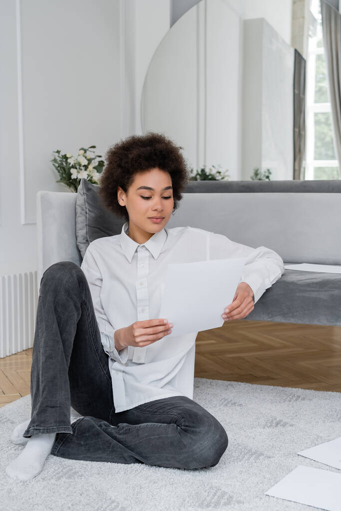 mujer afroamericana rizada sosteniendo documento en blanco mientras trabaja desde casa y sentado cerca de sofá de terciopelo - Foto, imagen