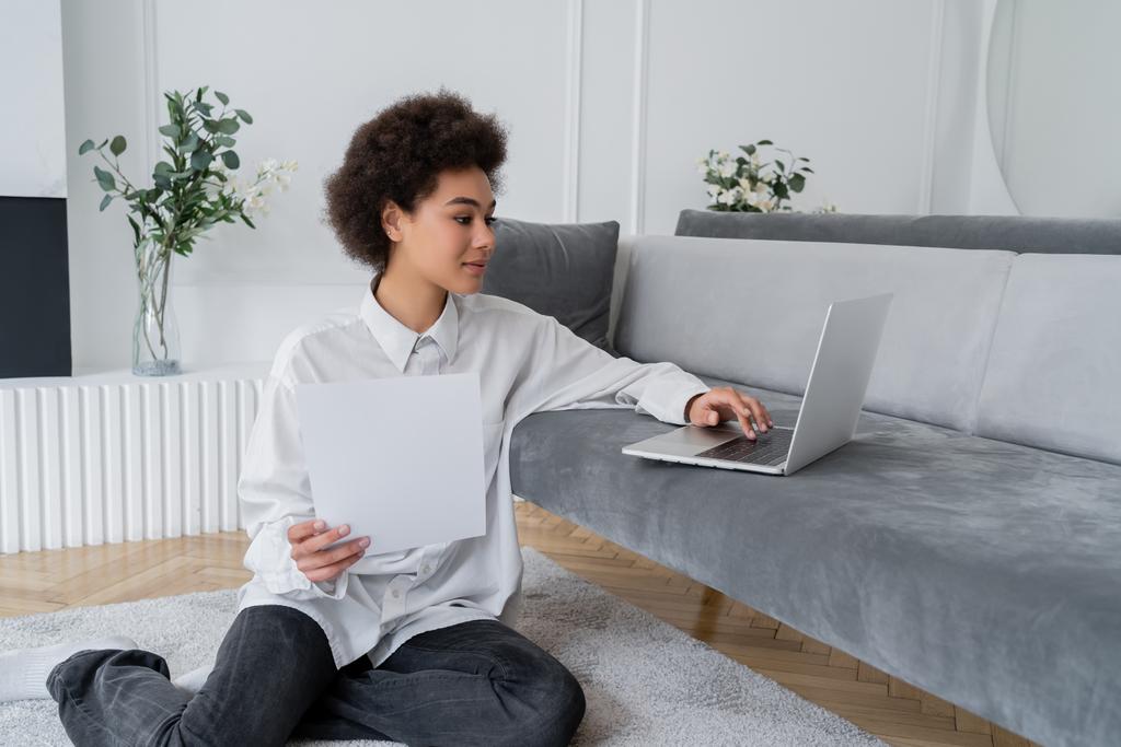 curly african american freelancer holding paper and using laptop on grey velvet sofa - Photo, Image