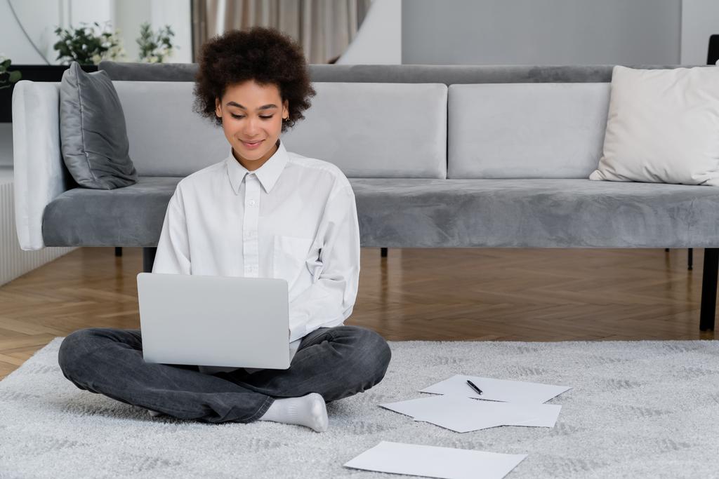 african american woman smiling while using laptop near documents on carpet  - Photo, Image