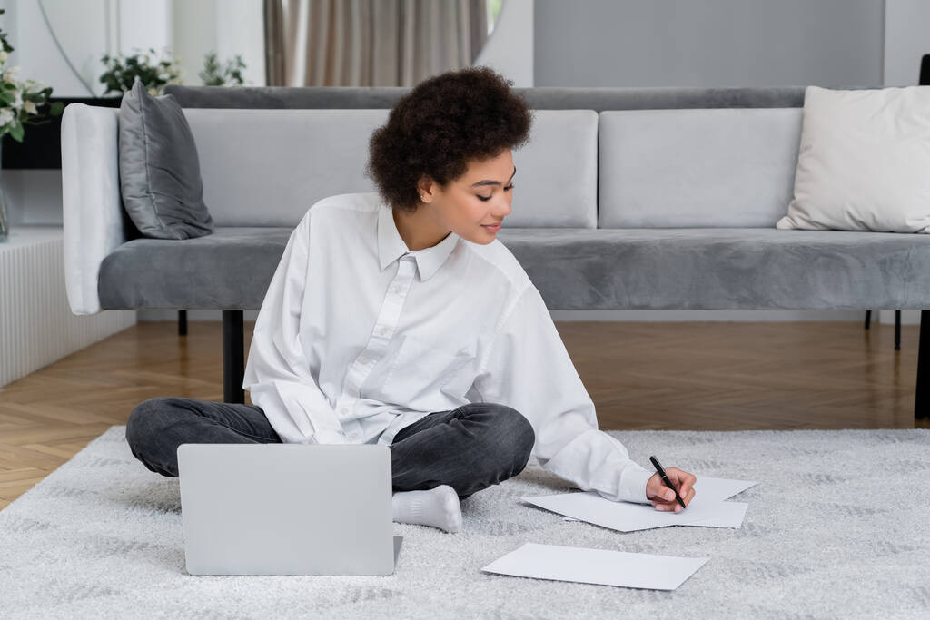 Afrikaans amerikaanse vrouw glimlachen tijdens het schrijven op papier en zitten met laptop op tapijt  - Foto, afbeelding