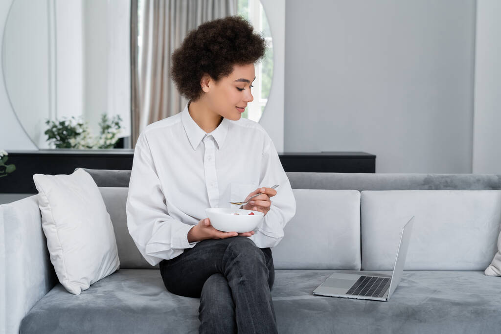 donna afroamericana che tiene la ciotola con la colazione mentre guarda il computer portatile sul divano di velluto - Foto, immagini