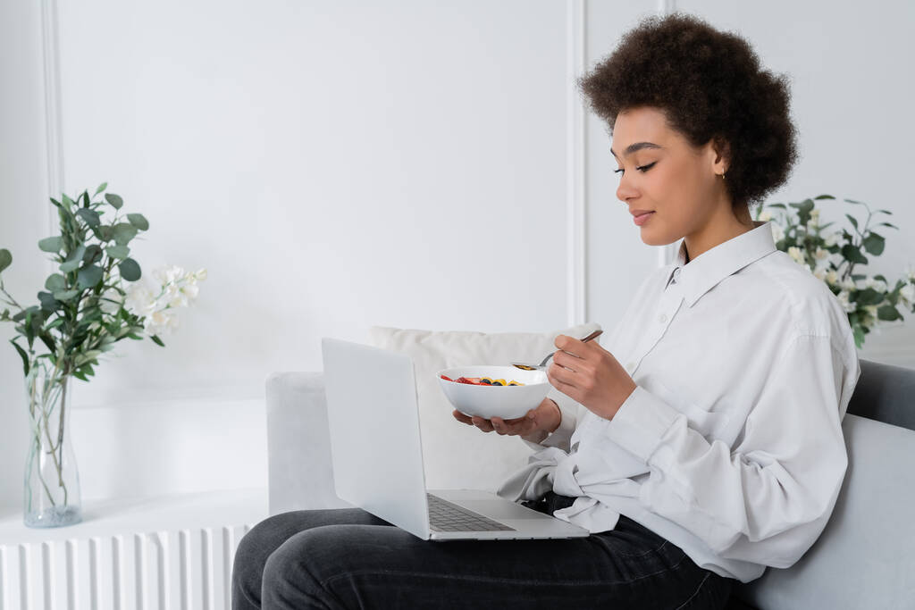 afro-américaine femme tenant bol avec des baies tout en regardant un film sur ordinateur portable et assis canapé en velours - Photo, image