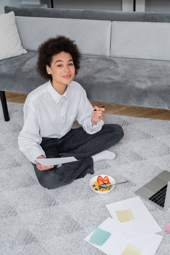 curly african american freelancer holding pen and paper while sitting on carpet near bowl with breakfast and laptop - Photo, Image