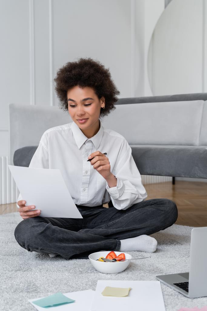 jovem Africano americano freelancer segurando caneta e papel enquanto sentado no tapete perto tigela com café da manhã e laptop - Foto, Imagem