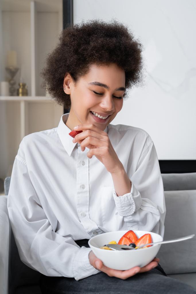 cheerful african american woman holding bowl with corn flakes and fresh strawberry - Photo, Image