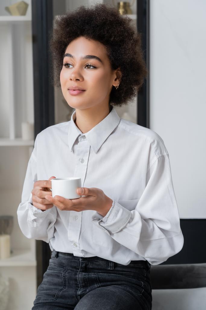 mujer afroamericana rizada en camisa blanca con cuello sosteniendo taza de café en casa - Foto, imagen