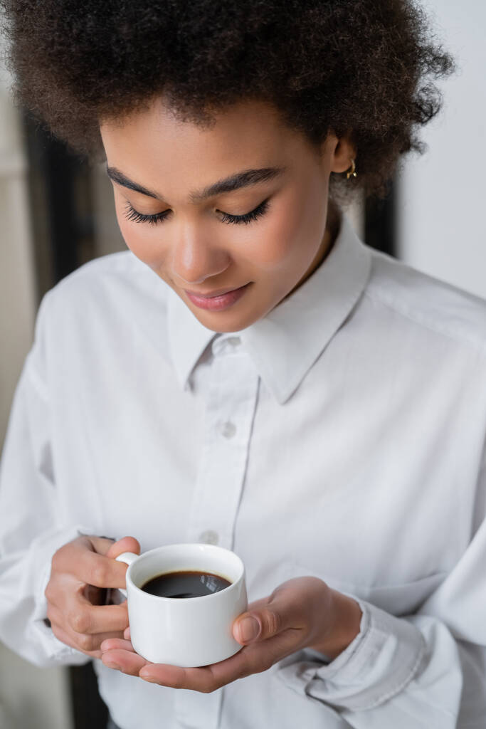 κατσαρομάλλη Αφροαμερικανή με λευκό πουκάμισο να κοιτάει καφέ.  - Φωτογραφία, εικόνα