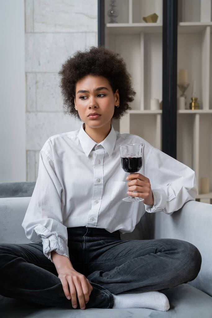 λυπημένη νεαρή Αφροαμερικανή που κρατάει ένα ποτήρι κόκκινο κρασί ενώ κάθεται σε βελούδινο καναπέ. - Φωτογραφία, εικόνα