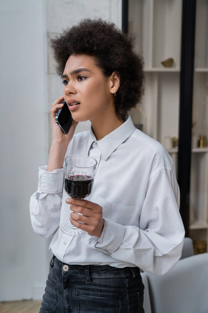 Unzufriedene Afroamerikanerin hält Glas Rotwein in der Hand und spricht auf Smartphone  - Foto, Bild