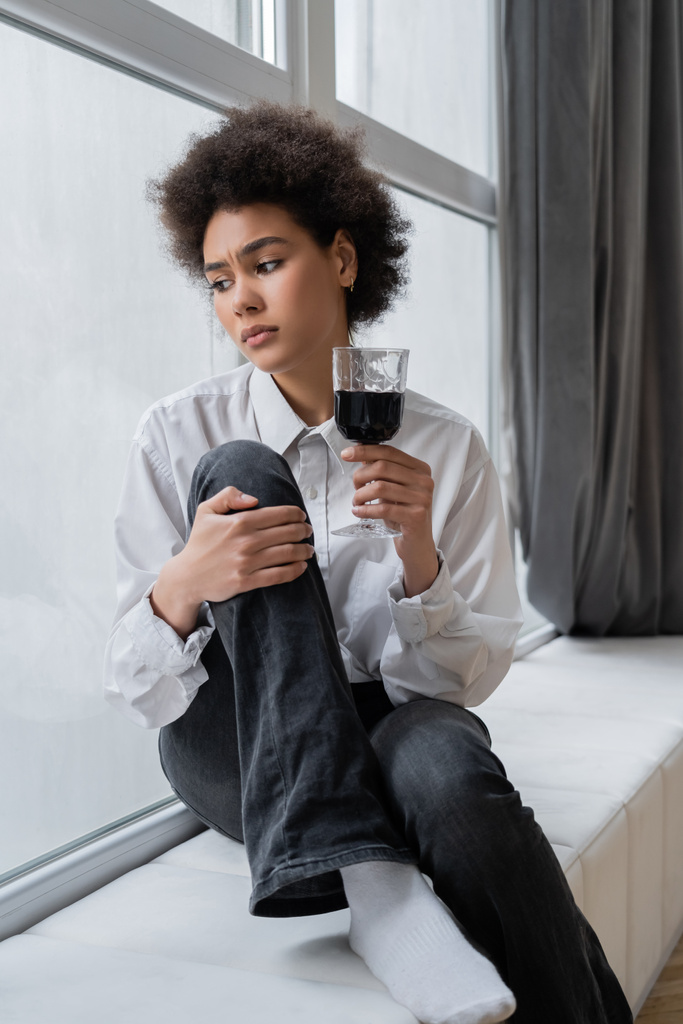 triste femme afro-américaine tenant un verre de vin rouge et regardant la fenêtre tout en étant assis au rebord de la fenêtre  - Photo, image