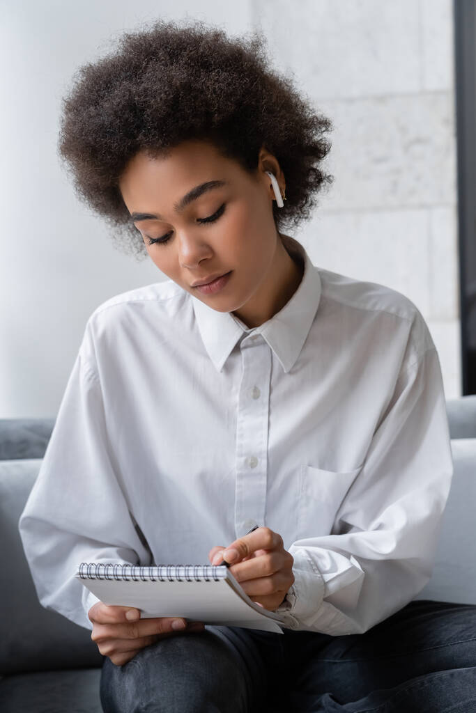 σγουρά αφροαμερικάνικη γυναίκα σε λευκό πουκάμισο και ασύρματο ακουστικό γράφοντας στο σημειωματάριο - Φωτογραφία, εικόνα