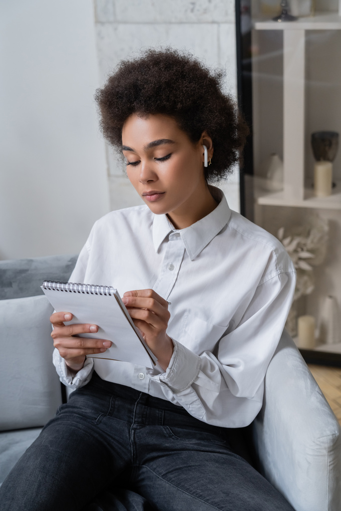 kręcone Afroamerykanka kobieta w białej koszuli i bezprzewodowe słuchawki robi notatki w notatniku - Zdjęcie, obraz