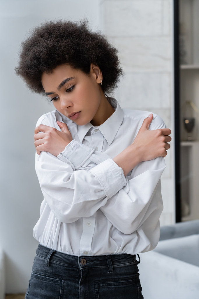 mujer afroamericana deprimida en camisa blanca abrazándose de hombros en casa - Foto, imagen