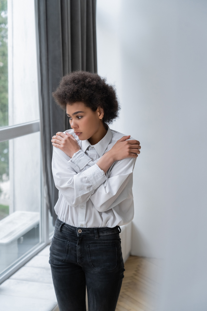 mulher afro-americana deprimida em camisa branca e jeans abraçando ombros em casa - Foto, Imagem