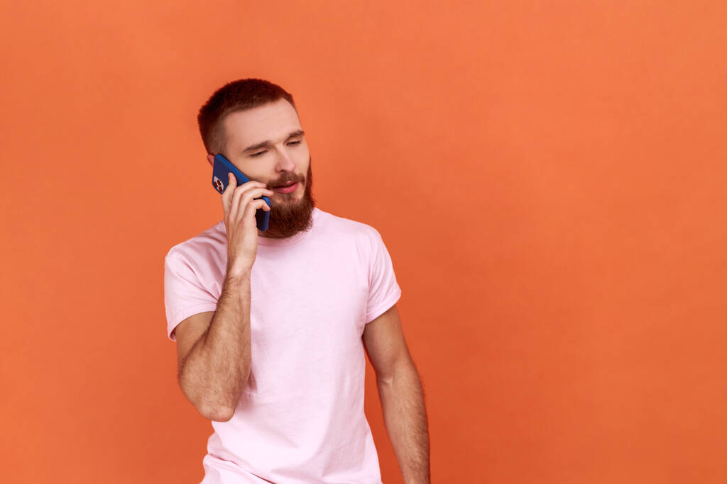 Portrait of handsome positive bearded man talking phone, calling to discuss plans, affordable tariffs, roaming, wearing pink T-shirt. Indoor studio shot isolated on orange background. - Photo, Image