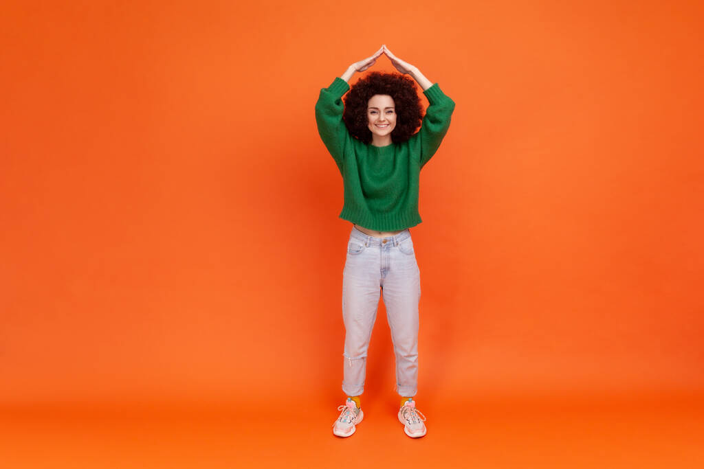 Full length portrait of woman with Afro hairstyle wearing green casual style sweater holding arms above head making roof gesture, insurance. Indoor studio shot isolated on orange background. - Photo, Image
