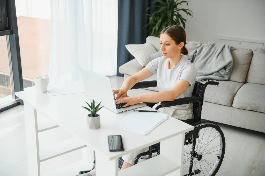 Freelancer en silla de ruedas con portátil cerca de notebook y papeles en la mesa. - Foto, Imagen