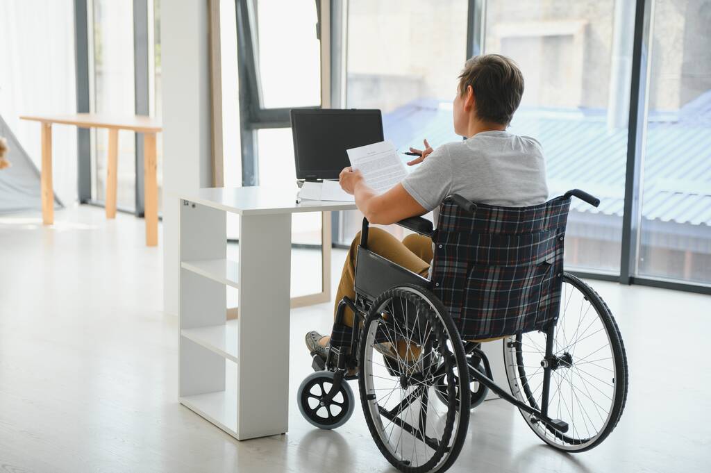 Kobieta w średnim wieku za pomocą laptopa siedzi na wózku inwalidzkim w domu. - Zdjęcie, obraz