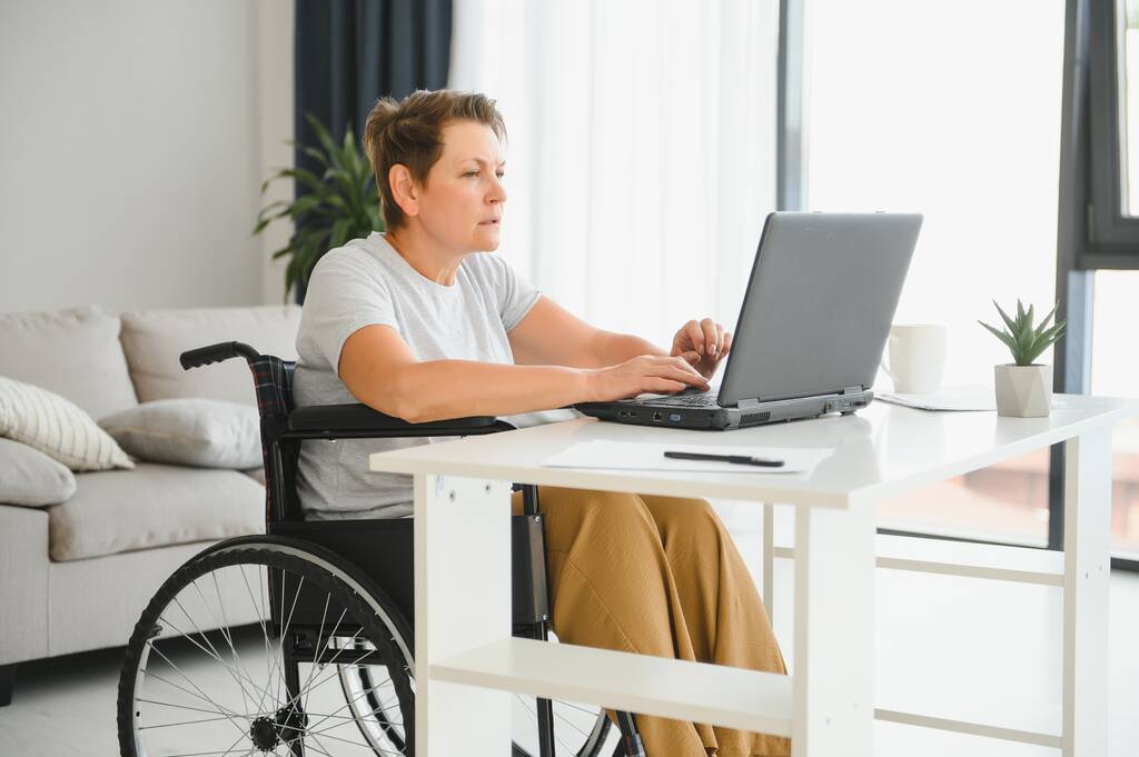 Freelance en fauteuil roulant en utilisant un ordinateur portable près d'un ordinateur portable et des papiers sur la table. - Photo, image