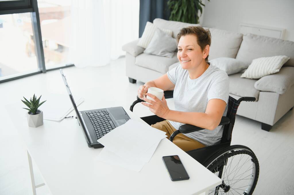 Ελεύθερος επαγγελματίας σε αναπηρική καρέκλα χρησιμοποιώντας φορητό υπολογιστή κοντά σε σημειωματάριο και χαρτιά στο τραπέζι. - Φωτογραφία, εικόνα