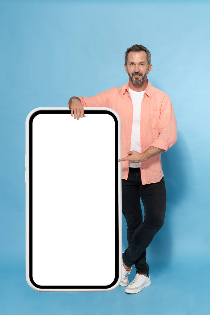 Middle aged grey haired man leaned on huge, giant smartphone pointing on white screen happy smiling on camera wearing peach shirt isolated on blue background. Mature fit man with smartphone. - Photo, Image