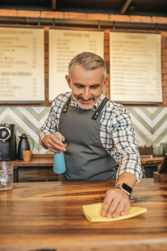 Wischfläche. Lächelnder Mann in Schürze spritzt Waschmittel und wischt Oberfläche der Theke im Café vor dem Hintergrund der Speisekarte - Foto, Bild