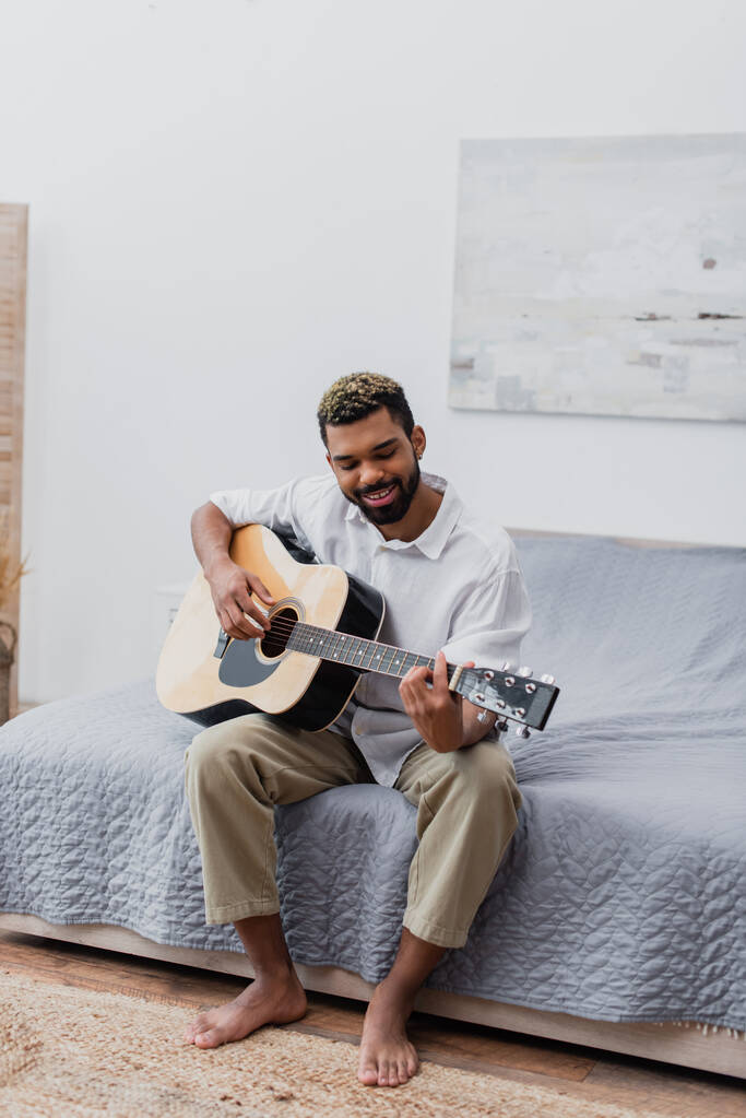 веселый молодой африканский американец с окрашенными волосами и бородой, играющий на акустической гитаре в спальне - Фото, изображение