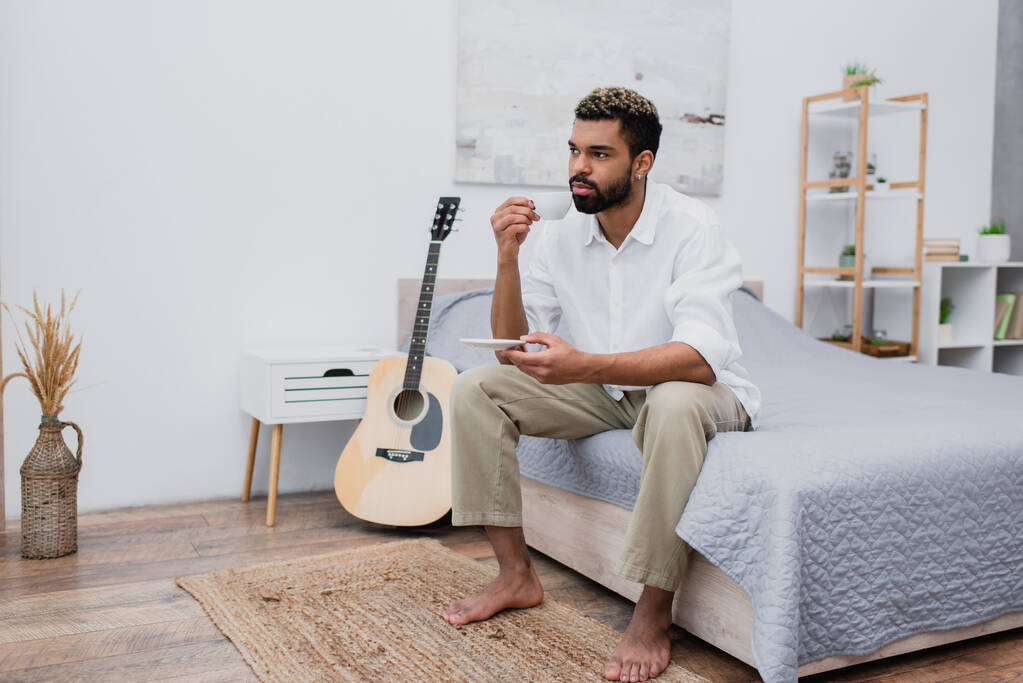 junger afrikanisch-amerikanischer Mann mit gefärbten Haaren, der eine Tasse Kaffee und eine Untertasse hält, während er auf dem Bett neben der Akustikgitarre sitzt - Foto, Bild