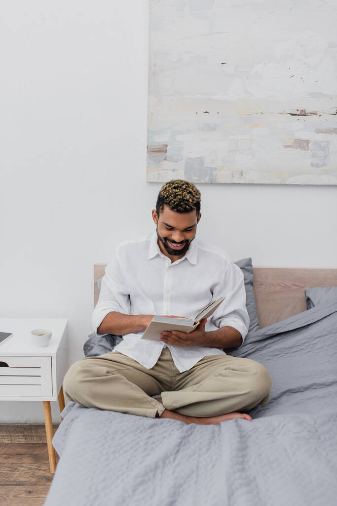 hombre afroamericano feliz con libro de lectura de pelo teñido en dormitorio moderno - Foto, imagen