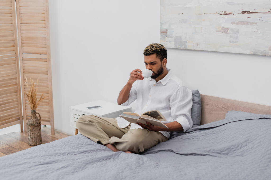 молодий афроамериканський чоловік з фарбованим волоссям читає книгу і п'є каву в сучасній спальні
 - Фото, зображення
