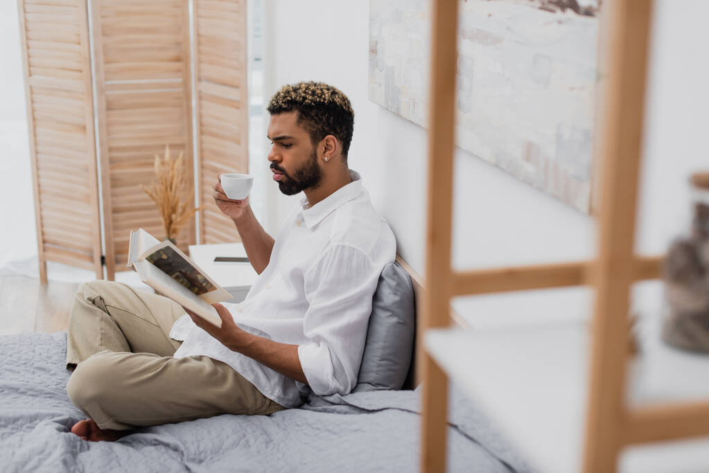 junger afrikanisch-amerikanischer Mann mit gefärbten Haaren liest Buch und hält Tasse Kaffee im modernen Schlafzimmer - Foto, Bild