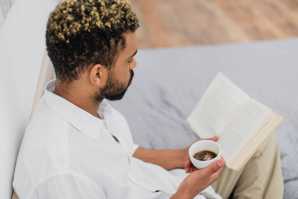 високий кут зору молодого афроамериканця з пофарбованим волоссям читаючи роман і тримаючи чашку кави в спальні
 - Фото, зображення