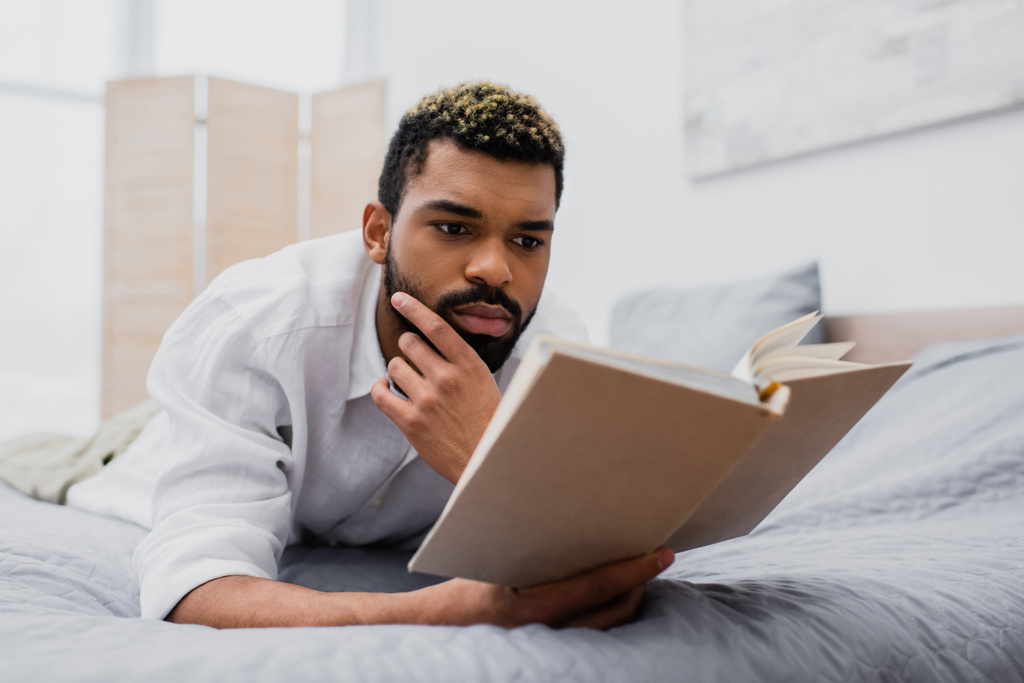 young african american man with dyed hair reading novel while lying on bed - Photo, Image