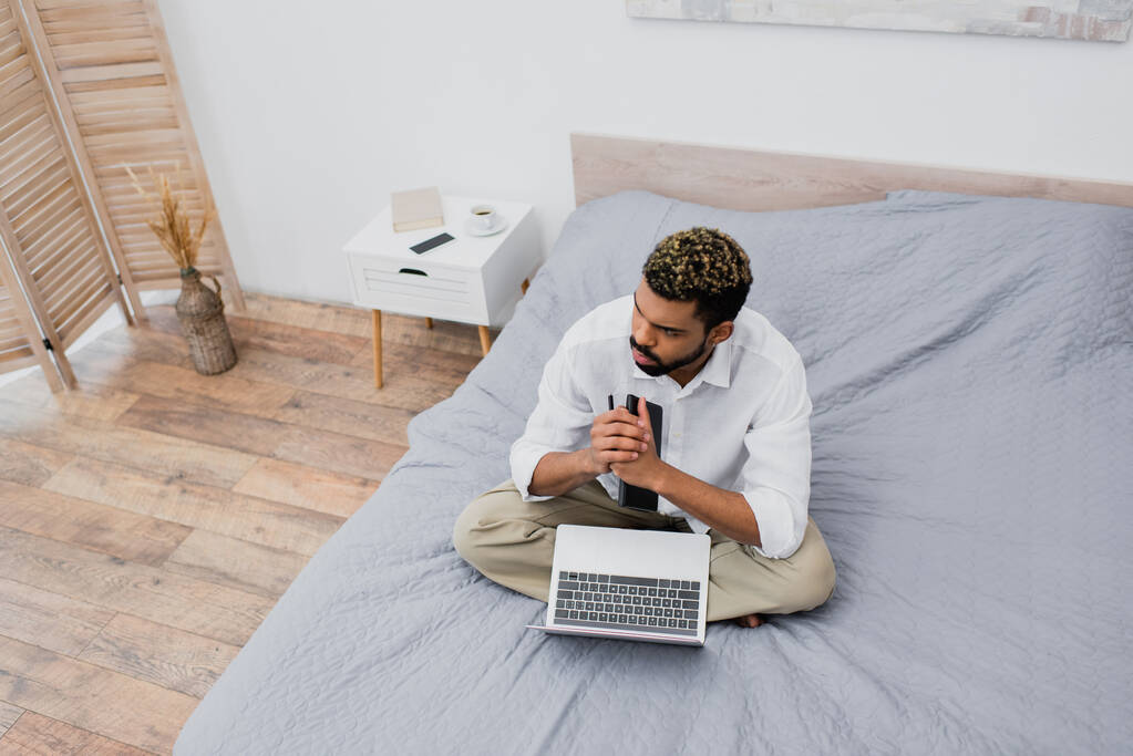 Hochwinkelaufnahme eines nachdenklichen afrikanisch-amerikanischen Mannes mit gefärbten Haaren, der mit Laptop auf dem Bett sitzt - Foto, Bild