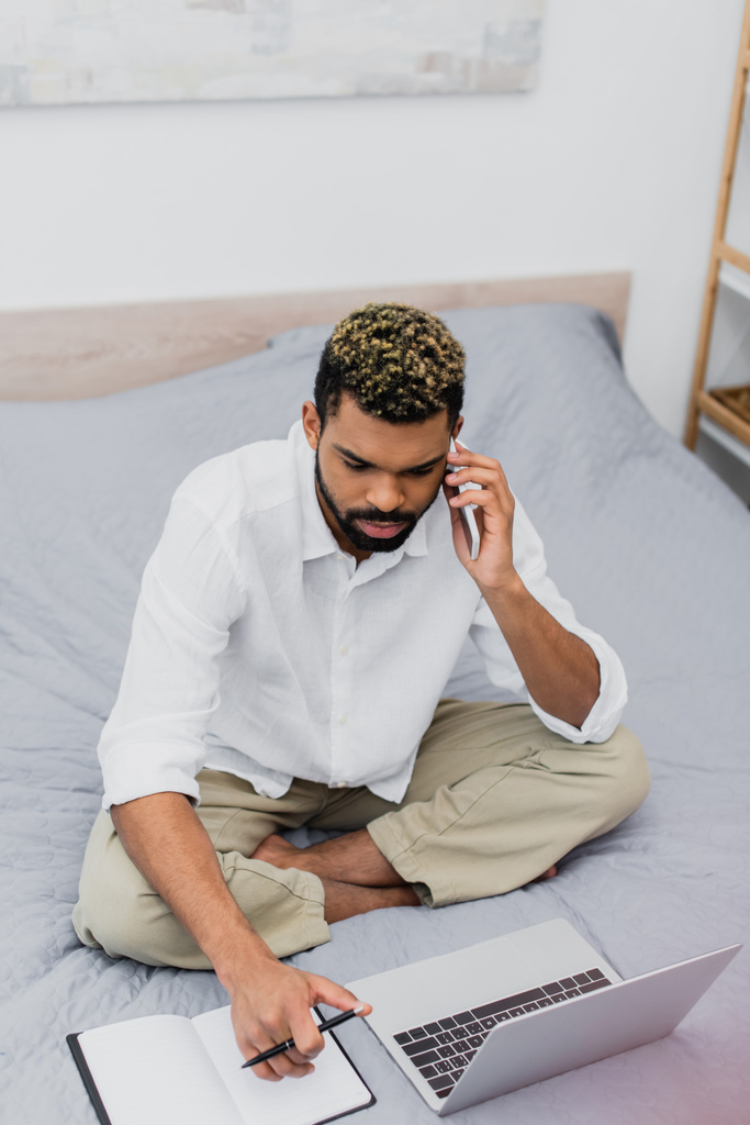 высокий угол зрения молодых африканских американец сидит на кровати во время разговора на смартфоне и с помощью ноутбука  - Фото, изображение