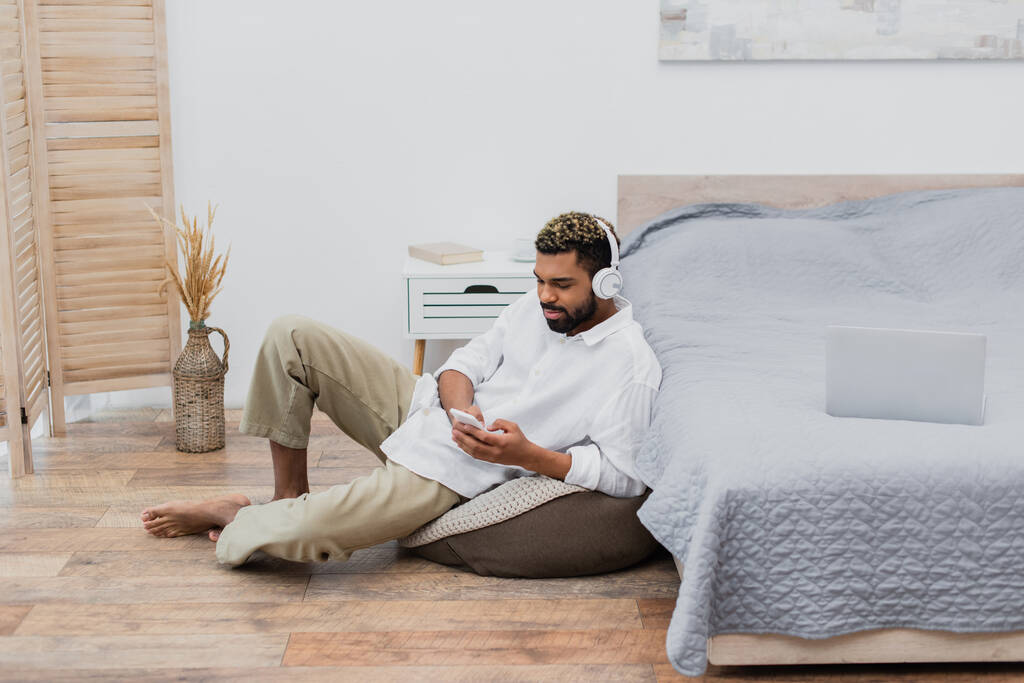 nuori afrikkalainen amerikkalainen mies langattomissa kuulokkeissa, joissa on älypuhelin, kun istut kannettavan tietokoneen vieressä sängyssä - Valokuva, kuva